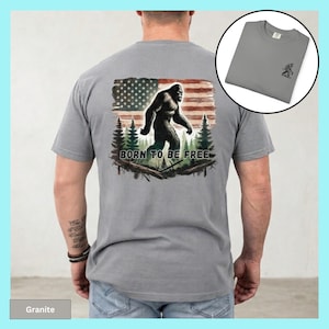 May include: A gray t-shirt featuring a Bigfoot graphic in front of an American flag and the text "BORN TO BE FREE". The shirt also has a small Bigfoot graphic on the upper left chest. The shirt is made of a soft material.