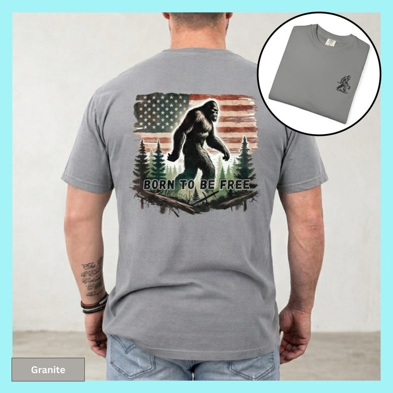 May include: A gray t-shirt featuring a Bigfoot graphic in front of an American flag and the text "BORN TO BE FREE". The shirt also has a small Bigfoot graphic on the upper left chest. The shirt is made of a soft material.