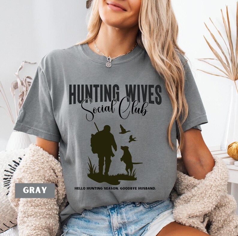 K&ouml;nnte beinhalten: Graues T-Shirt mit dem Text "Hunting Wives Social Club" in Schwarz. Das Shirt zeigt eine Silhouette eines J&auml;gers mit einem Hund und V&ouml;geln. Der Text "Hello hunting season. Goodbye husband." ist unter dem Bild gedruckt.