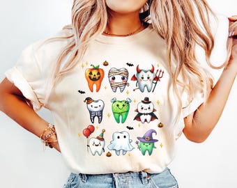 May include: A white t-shirt with a Halloween-themed design featuring cartoon teeth dressed in costumes. The teeth are wearing costumes such as a pumpkin, a mummy, a vampire, a ghost, and a witch. The shirt has a festive and fun design.