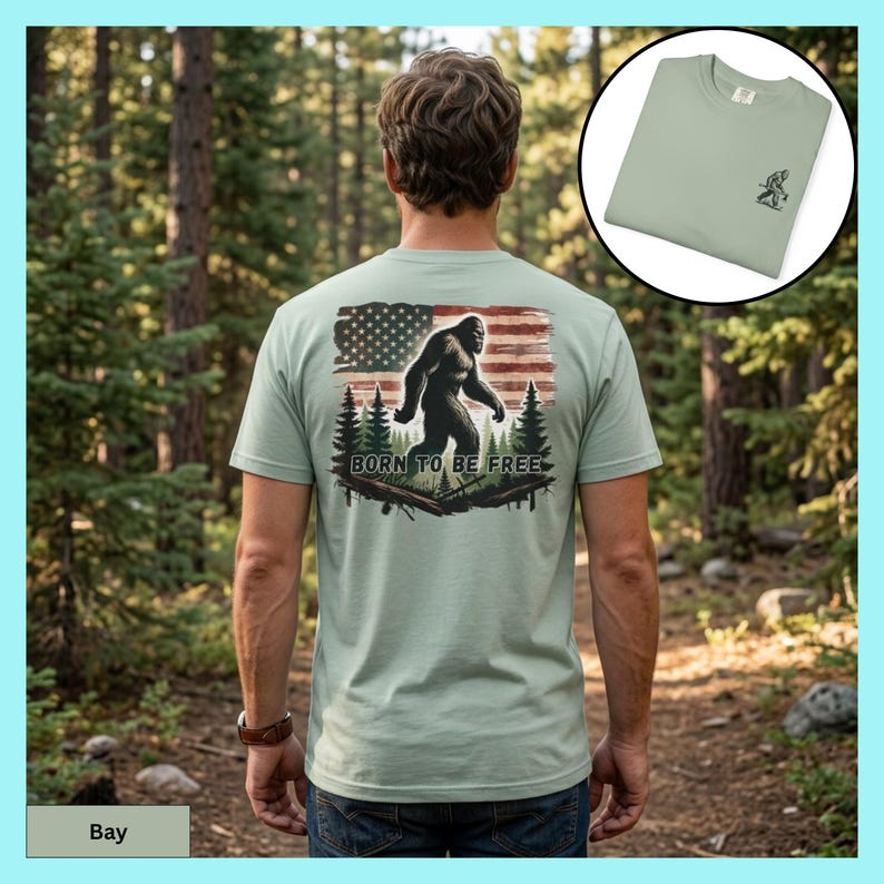 May include: Sage green t-shirt featuring a Bigfoot graphic with an American flag and the text "Born to be Free." The shirt is displayed on a person in a forest setting. A smaller image of the shirt shows a Bigfoot graphic on the chest.