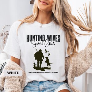 K&ouml;nnte beinhalten: Wei&szlig;es T-Shirt mit einer schwarzen Grafik, auf der "Hunting Wives Social Club" steht. Die Grafik zeigt auch eine Silhouette eines J&auml;gers mit einem Hund und V&ouml;geln. Der Text unter der Grafik lautet "Hello hunting season. Goodbye husband."