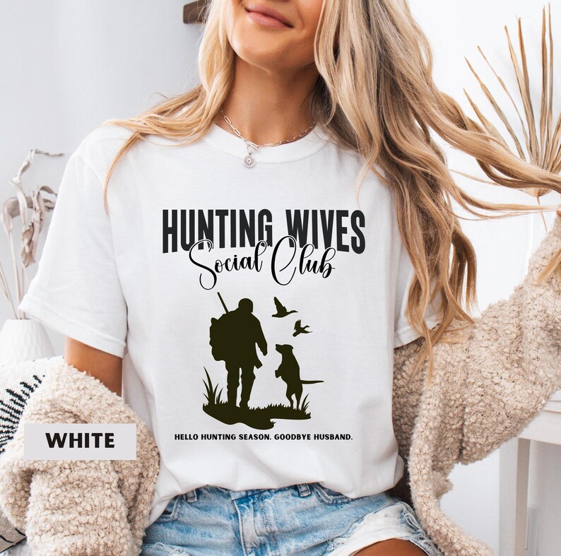 K&ouml;nnte beinhalten: Wei&szlig;es T-Shirt mit einer schwarzen Grafik, auf der "Hunting Wives Social Club" steht. Die Grafik zeigt auch eine Silhouette eines J&auml;gers mit einem Hund und V&ouml;geln. Der Text unter der Grafik lautet "Hello hunting season. Goodbye husband."