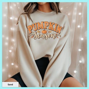 May include: A sand-colored crewneck sweatshirt featuring the text PUMPKIN SEASON in orange and brown block and script lettering. The design includes a central orange pumpkin illustration surrounded by small autumn leaves and foliage accents.