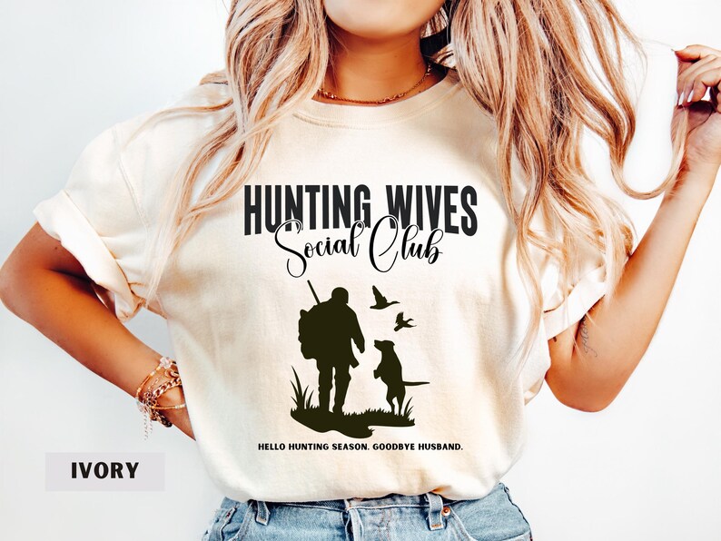 K&ouml;nnte beinhalten: Ein T-Shirt in Elfenbein mit dem Text "Hunting Wives Social Club" und einer Silhouette eines J&auml;gers mit einem Hund und V&ouml;geln. Der Text unter der Grafik lautet "Hello hunting season, goodbye husband."