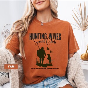 K&ouml;nnte beinhalten: Ein orangefarbenes T-Shirt mit dem Text "Hunting Wives Social Club" in Schwarz. Unter dem Text befindet sich eine Silhouette eines J&auml;gers mit einem Hund. Der Text "Hello Hunting Season, Goodbye Husband." befindet sich am unteren Rand des Shirts.