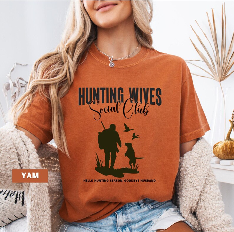 K&ouml;nnte beinhalten: Ein orangefarbenes T-Shirt mit dem Text "Hunting Wives Social Club" in Schwarz. Unter dem Text befindet sich eine Silhouette eines J&auml;gers mit einem Hund. Der Text "Hello Hunting Season, Goodbye Husband." befindet sich am unteren Rand des Shirts.