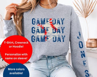 May include: A heather gray crewneck sweatshirt featuring the text GAME DAY repeated three times in a distressed blue font, overlaid with a red baseball stitch graphic. The left sleeve displays the name JONES printed vertically in blue.