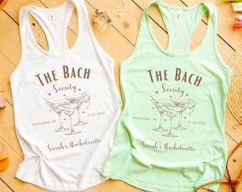 May include: Two white and light green tank tops with the text "The Bach Society" and a drawing of three martini glasses. The text also includes "Scottsdale, AZ" and "5-18-2024". The text "Sarah's Bachelorette" is at the bottom of each tank top.