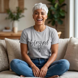 May include: A heather grey short-sleeved t-shirt featuring the word blessed in a black cursive script font across the chest, positioned above a simple black hand-drawn heart outline. The shirt has a crew neckline and a standard fit.