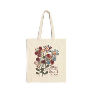 Peut inclure: Un sac fourre-tout en toile beige orné d'un motif de cinq fleurs stylisées aux motifs variés à carreaux et à pois dans des tons de rouge, bleu et marron. Le texte "Jesus loves me, this I know" est imprimé en marron à côté des tiges.