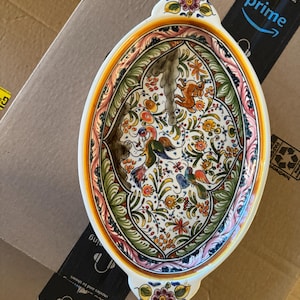 May include: An oval ceramic serving dish with two handles, featuring a white background decorated with colorful hand-painted floral patterns, green foliage, and small animal figures in shades of orange, blue, yellow, and pink, with a yellow rim.