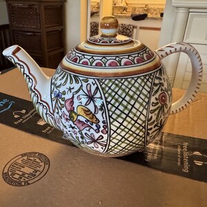May include: A ceramic teapot featuring a hand-painted design with a green lattice pattern, floral motifs, and small bird illustrations in shades of yellow, pink, and green. The teapot has a matching lid with a knob and a curved handle and spout.