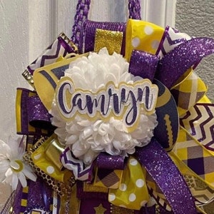 Op de afbeelding: Een paars-gouden cheer-boog met een witte pompon en een naamsticker met de tekst "Camryn".