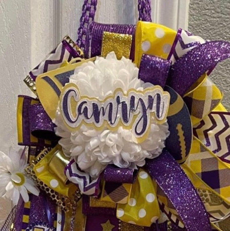 Op de afbeelding: Een paars-gouden cheer-boog met een witte pompon en een naamsticker met de tekst "Camryn".