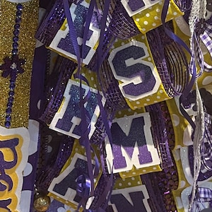 May include: Purple and gold graduation door swag with the letters "H.R.S.A.M.N." and a heart-shaped sign that says "To Mom & Dad Avery".