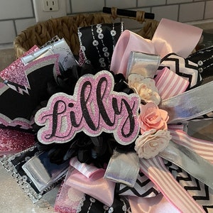 Op de afbeelding: Een roze en zwarte strik met een glinsterend roze bord waarop in zwarte letters "Lilly" staat. De strik is gemaakt van verschillende linten, waaronder roze, zwart, wit en zilver. Er zitten ook wat witte en roze bloemen op de strik.