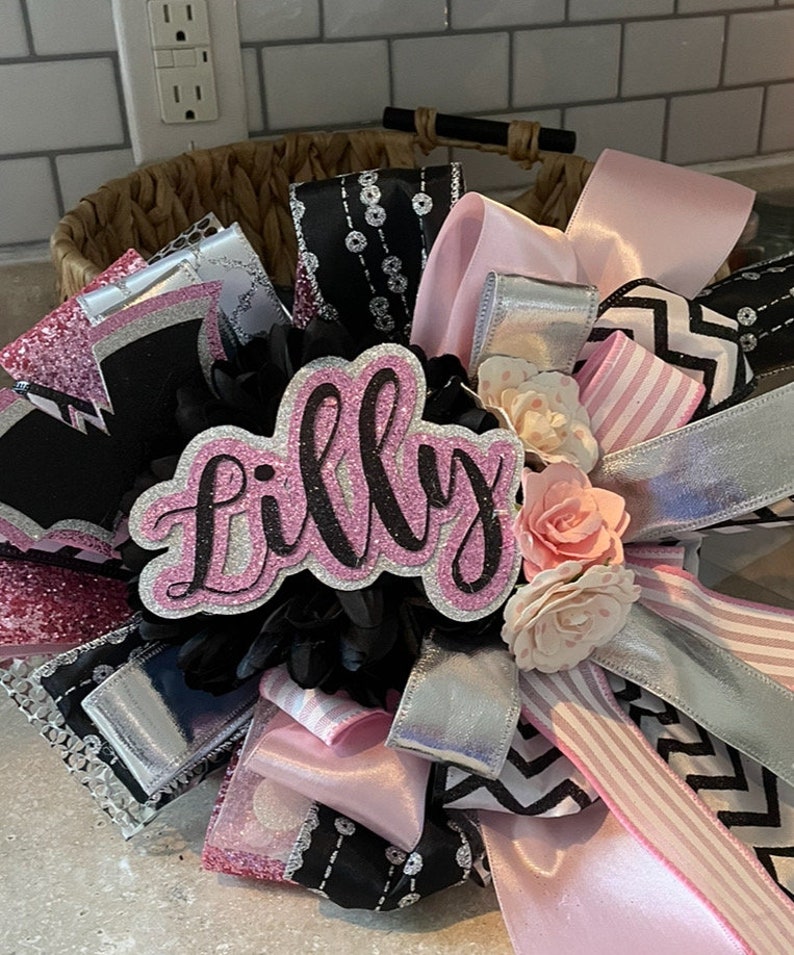 Op de afbeelding: Een roze en zwarte strik met een glinsterend roze bord waarop in zwarte letters "Lilly" staat. De strik is gemaakt van verschillende linten, waaronder roze, zwart, wit en zilver. Er zitten ook wat witte en roze bloemen op de strik.