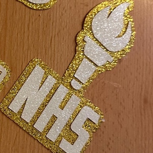 May include: A gold and white glitter NHS emblem featuring a torch design. The letters "NHS" are in white, outlined with gold glitter. The torch is also white with gold glitter accents. This is likely a decorative item.