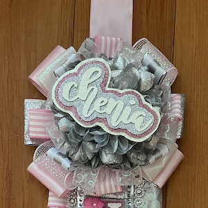 Op de afbeelding: Een roze en zilveren afstudeersjerp met het woord "Chelsea" geschreven in zilveren glitterletters op een roze achtergrond.