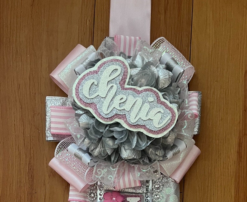 Op de afbeelding: Een roze en zilveren afstudeersjerp met het woord "Chelsea" geschreven in zilveren glitterletters op een roze achtergrond.