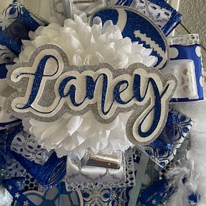 Op de afbeelding: Een witte en blauwe cheer bow met zilveren glitteraccenten. De strik heeft een witte bloem, een blauwe en zilveren voetbal en een zilveren glitternaamkaartje met de tekst "Laney".