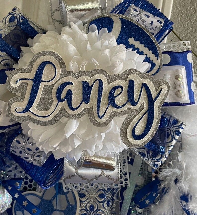 Op de afbeelding: Een witte en blauwe cheer bow met zilveren glitteraccenten. De strik heeft een witte bloem, een blauwe en zilveren voetbal en een zilveren glitternaamkaartje met de tekst "Laney".