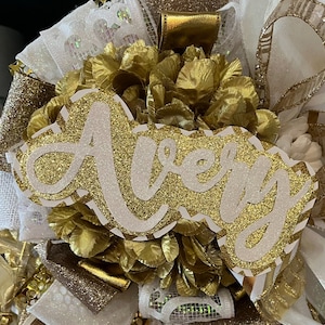 Op de afbeelding: Een wit en gouden glitternaam bord met de naam "Avery" in witte cursieve letters. Het bord is omringd door gouden en witte bloemen.