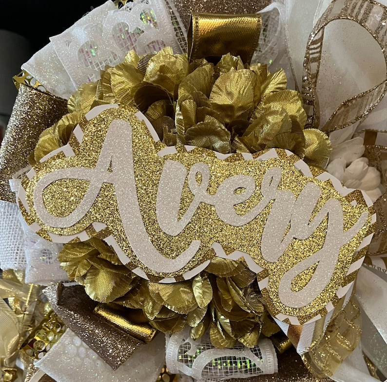 Op de afbeelding: Een wit en gouden glitternaam bord met de naam "Avery" in witte cursieve letters. Het bord is omringd door gouden en witte bloemen.