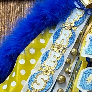 May include: A blue and gold graduation sash with the word "FRESHMAN" spelled out in white letters with gold trim. The sash is decorated with white flowers, gold beads, and blue and white polka dot ribbon.