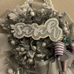 Op de afbeelding: Een zilveren en witte afstudeerlei met de naam "Sarah" gespeld in zilveren glitterletters.