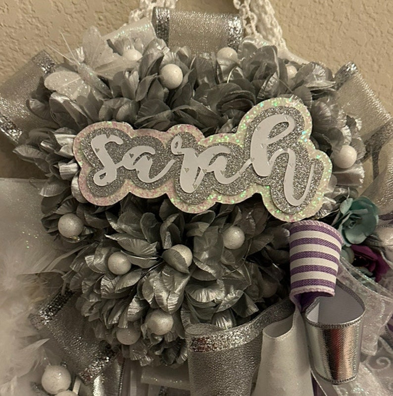 Op de afbeelding: Een zilveren en witte afstudeerlei met de naam "Sarah" gespeld in zilveren glitterletters.