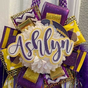 Op de afbeelding: Een paars-gouden afstudeerkrans met de naam "Ashlyn" in witte en paarse glitterletters.