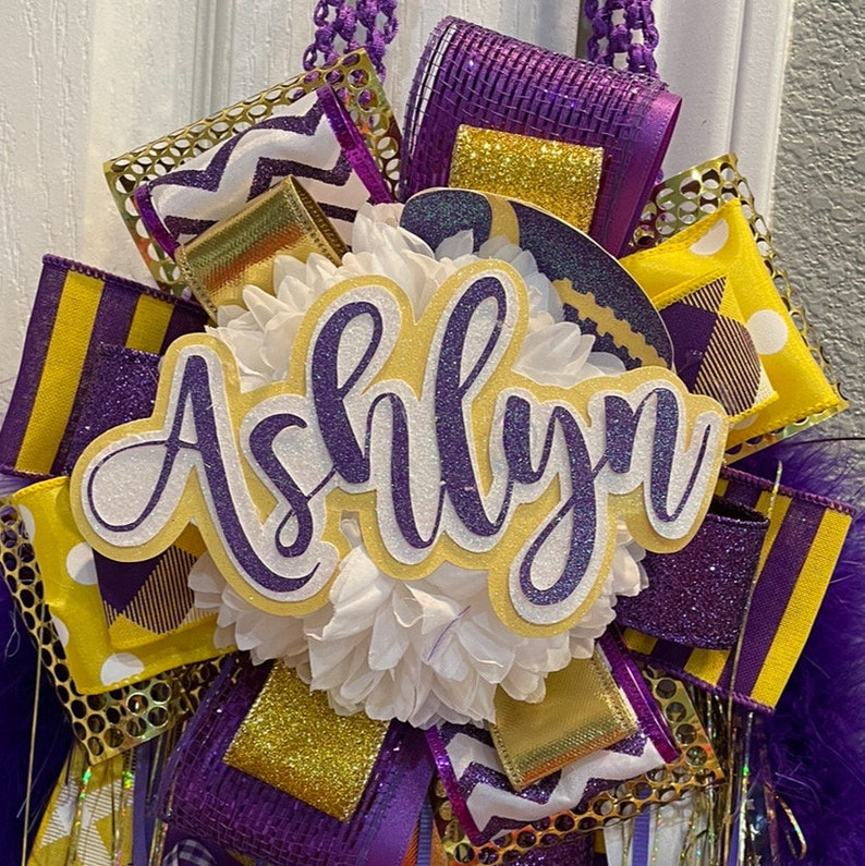 Op de afbeelding: Een paars-gouden afstudeerkrans met de naam "Ashlyn" in witte en paarse glitterletters.