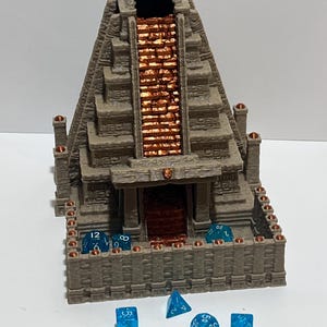May include: A detailed, miniature model of a Mesoamerican pyramid with a copper-colored staircase and accents. The model is gray with copper highlights and is surrounded by blue dice, perfect for tabletop gaming.