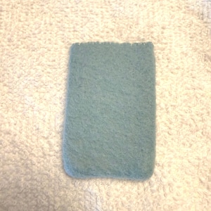 May include: A rectangular, light blue felt item with a textured surface. The edges are slightly rounded, and the top edge has a scalloped design. The item is set against a white, textured background.