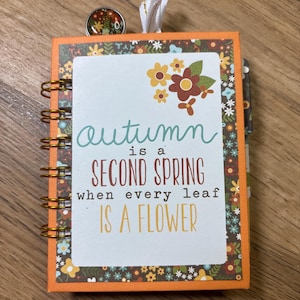 May include: A small spiral-bound notebook with an orange cover and a floral border. The center features the text: autumn is a SECOND SPRING when every leaf IS A FLOWER. A small circular charm with a floral pattern and a white ribbon are attached.