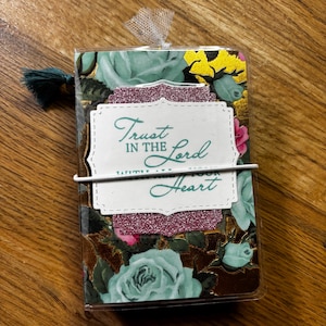 May include: A small rectangular notebook featuring a floral pattern of mint green roses and gold accents on a dark background. A white label with the text Trust IN THE Lord WITH ALL YOUR Heart is centered, accented by a glittery pink scalloped border.