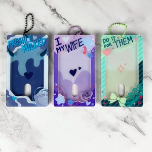 May include: Three rectangular plastic ID badge holders with ball chain attachments. The left holder is blue with the text "FRESHLY MILKED" and a bucket graphic. The center holder is purple with "I MY WIFE" text. The right holder is green with "DO IT FOR THEM" text.
