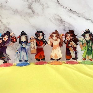 以下が含まれることがあります： カラフルなアニメキャラクターのアクリルスタンドが7つ。キャラクターはそれぞれ異なる衣装を着て、さまざまな小道具を持っています。スタンドは黄色の表面に置かれています。