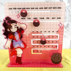 May include: A pink acrylic calendar with a grid of squares for each day of the month. The calendar has a cartoon character illustration of a girl with long brown hair wearing a red dress and white apron. The calendar has black and brown cartoon animal illustrations on the days of the month. The calendar has the months of the year printed across the top: JAN FEB MAR APR MAY JUN JUL AUG SEP OCT NOV DEC. The calendar has the days of the week printed across the top: MON TUE WED THUR FRI SAT SUN.