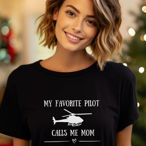 May include: Black crew neck t-shirt featuring the white text "MY FAVORITE PILOT CALLS ME MOM" and a helicopter graphic. A simple, heart-shaped symbol is below the text.