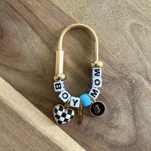 May include: A gold-toned keychain with the words "BOY" and "MOM" spelled out in white letter beads. It features a black and white checkered heart charm, a lightning bolt charm, a turquoise bead, and a black circular charm with the letter "J".