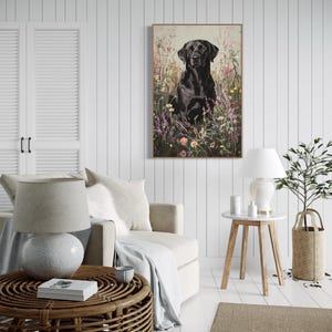 Pode incluir: Um labrador retriever preto sentado em um campo de flores silvestres. O c&atilde;o est&aacute; olhando diretamente para a c&acirc;mera. A pintura &eacute; emoldurada e pendurada em uma parede branca em uma sala de estar.