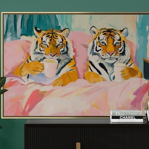 May include: A framed art print depicting two tigers in bed, each holding a cup. The tigers have orange and black stripes, set against a pink and white bedding background. The artwork is displayed above a black cabinet with decorative vases.