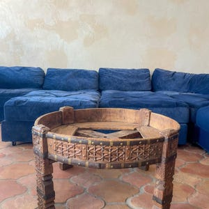 May include: A rustic, round wooden coffee table with intricate carved details and metal accents. The table has a distressed finish, showcasing its aged appearance. The legs are sturdy and carved, adding to the table's unique design. The table is approximately 24 inches in diameter.