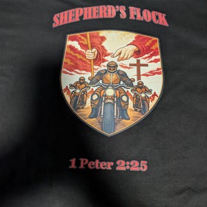 May include: A black t-shirt featuring a graphic print with the text SHEPHERD'S FLOCK at the top and 1 Peter 2:25 at the bottom. The central shield-shaped image depicts three figures in medieval armor riding motorcycles under a divine hand.
