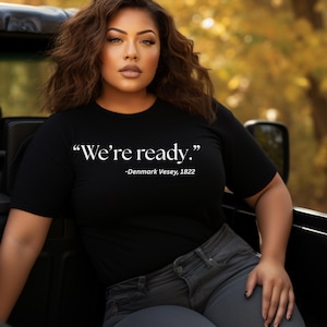 Può includere: Una donna indossa una maglietta nera con il testo "We're ready." -Denmark Vesey, 1822. È seduta in un veicolo con uno sfondo sfocato di foglie autunnali.
