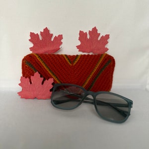 May include: A red, orange, and green crocheted glasses case with a pair of gray sunglasses on a white surface.  The case is decorated with three red maple leaves.