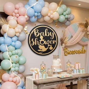 Puede incluir: Una decoración para baby shower con un gran letrero circular negro con el texto dorado Baby Shower, un icono de chupete y una cigüeña. El conjunto incluye un arco de globos en tonos pastel, detalles dorados de luna y estrella, y una mesa con pastel.